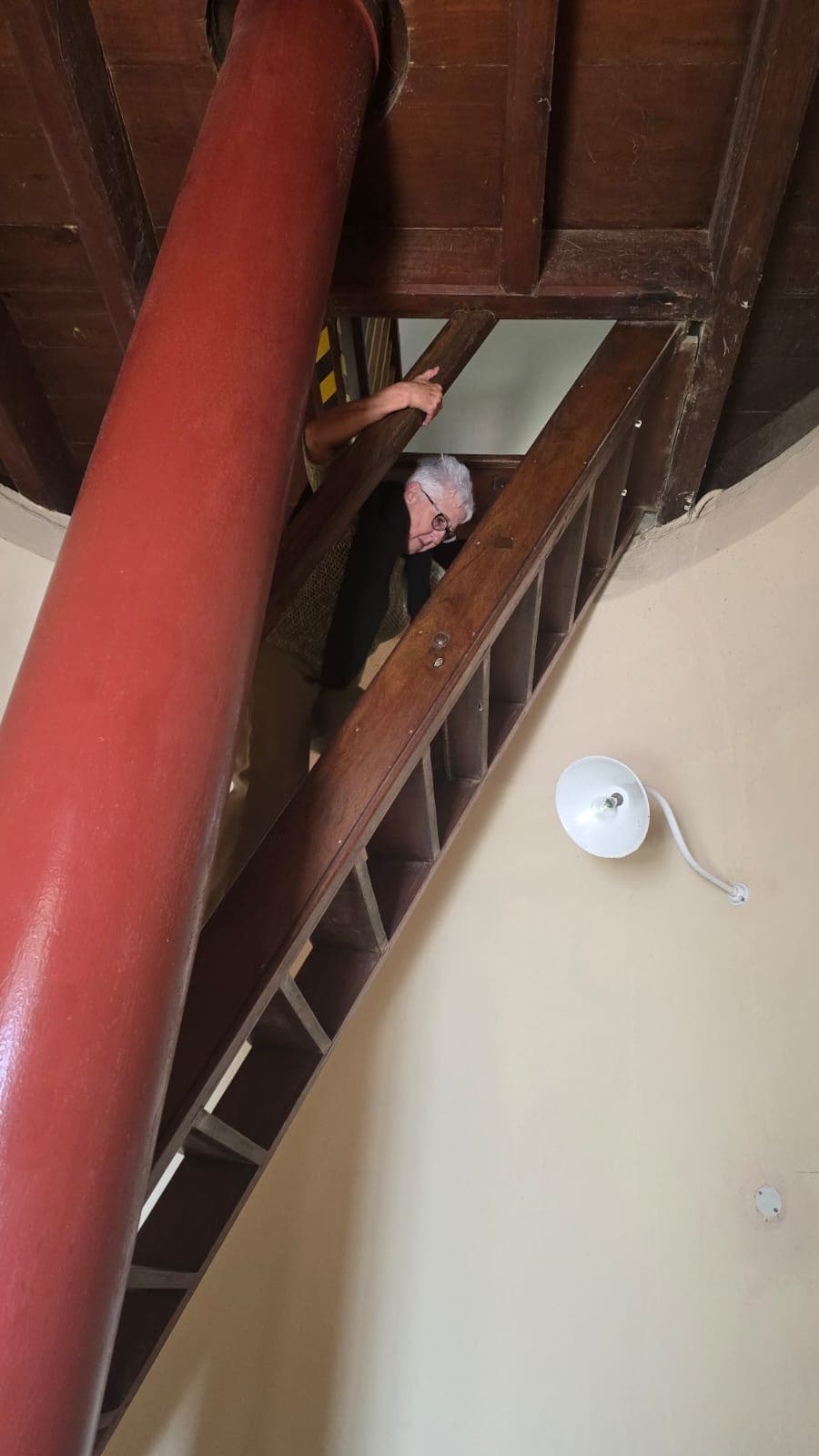 A person climbs a steep wooden ladder through a ceiling opening beside a large red pillar.