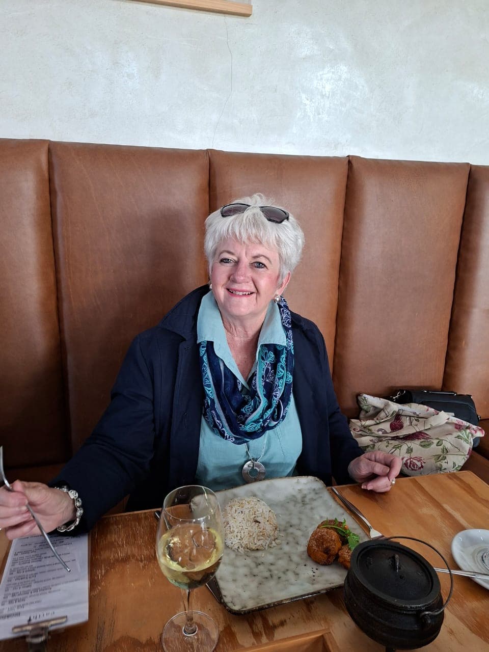 Smiling woman with short white hair sits at a restaurant table with rice and wine.