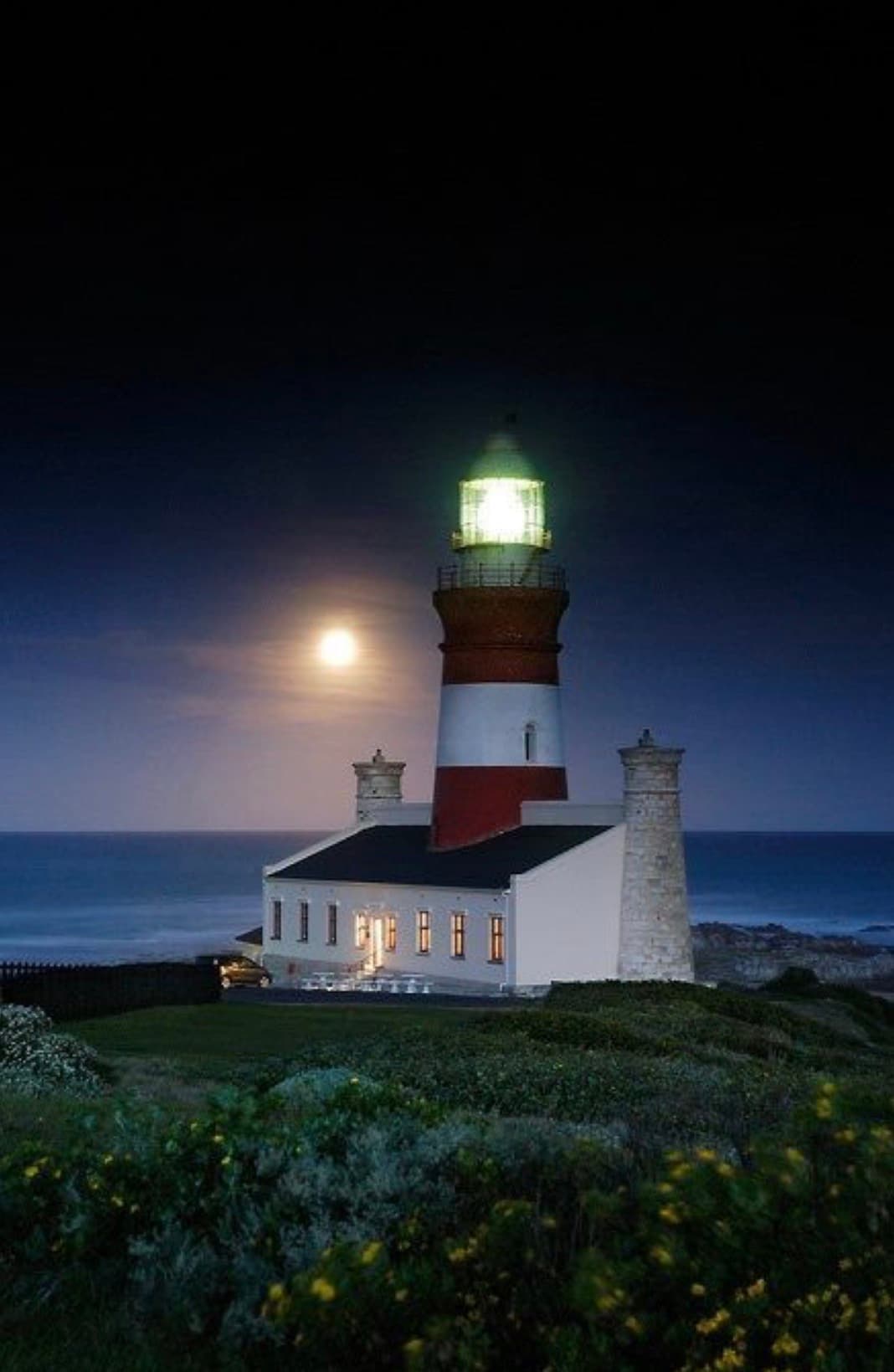 Red and white striped lighthouse glowing at night under a full moon beside the ocean.