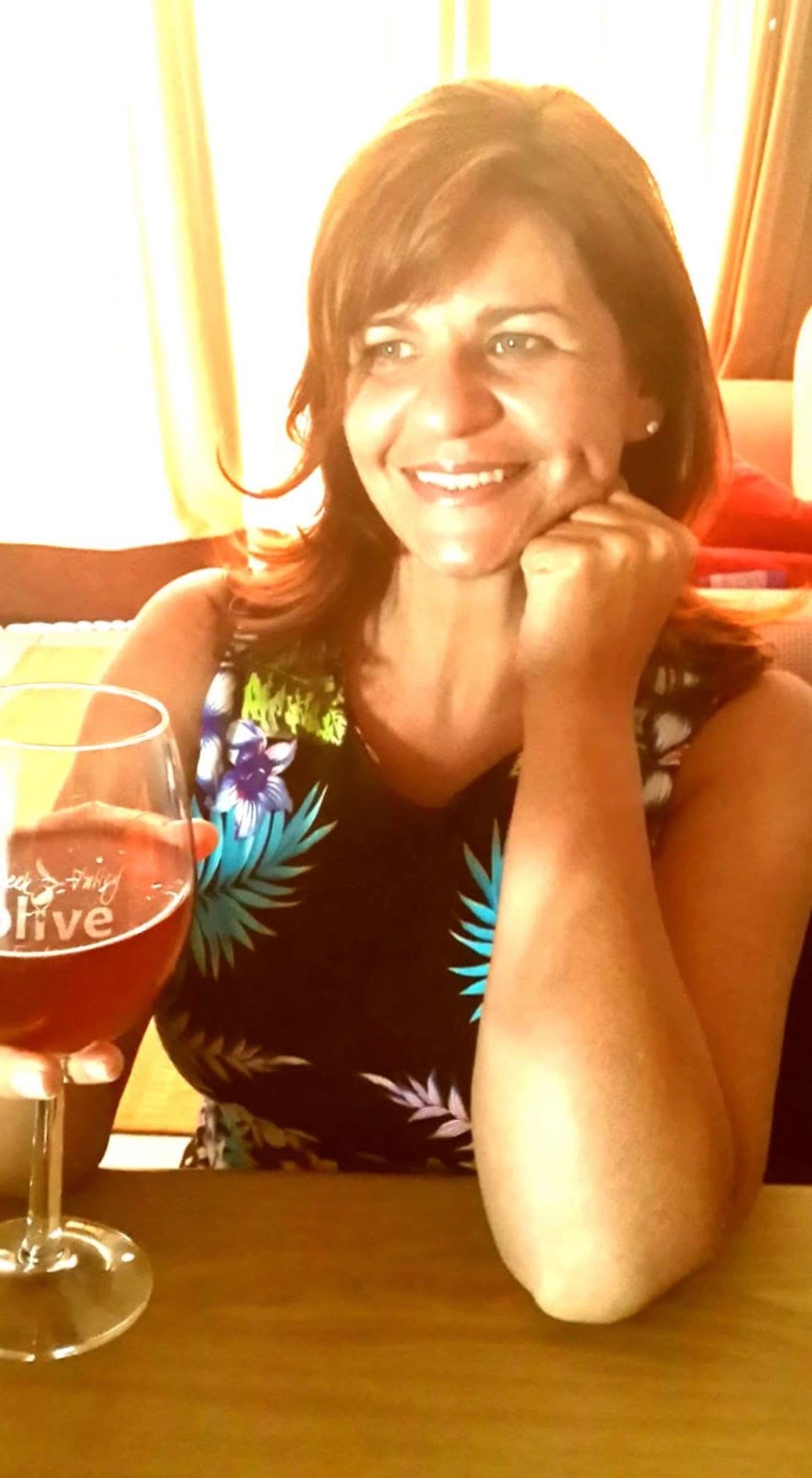 Smiling woman with auburn hair holding a wine glass, wearing a tropical floral top.