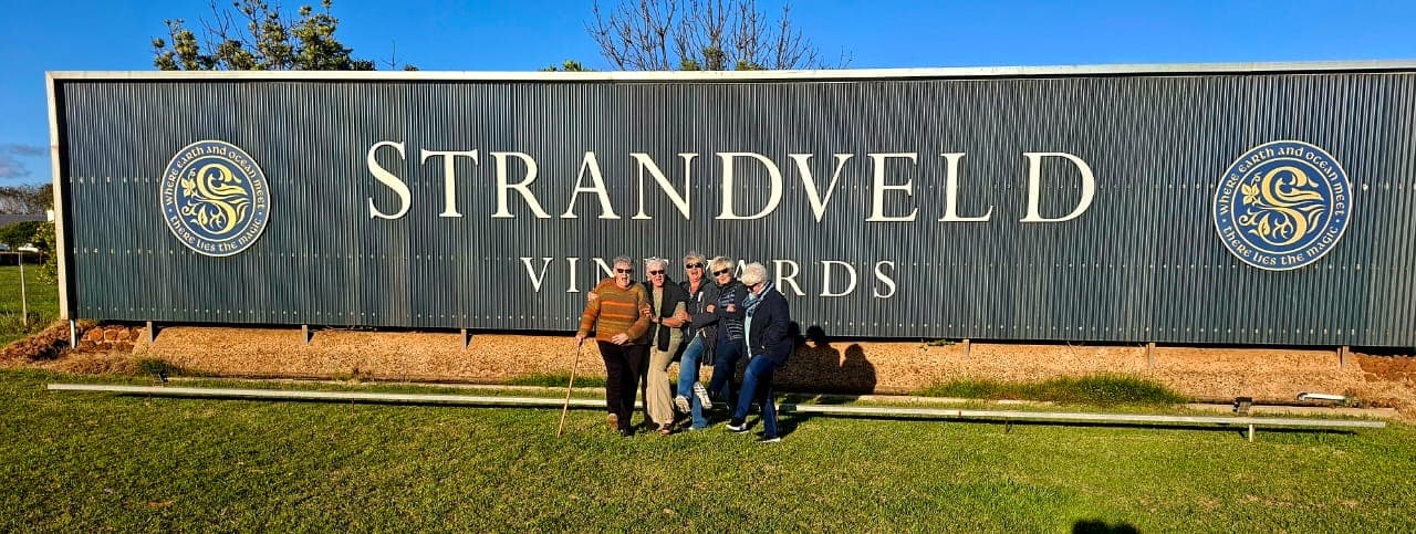 Five women pose happily in front of a large Strandveld Vineyards sign on a lawn.