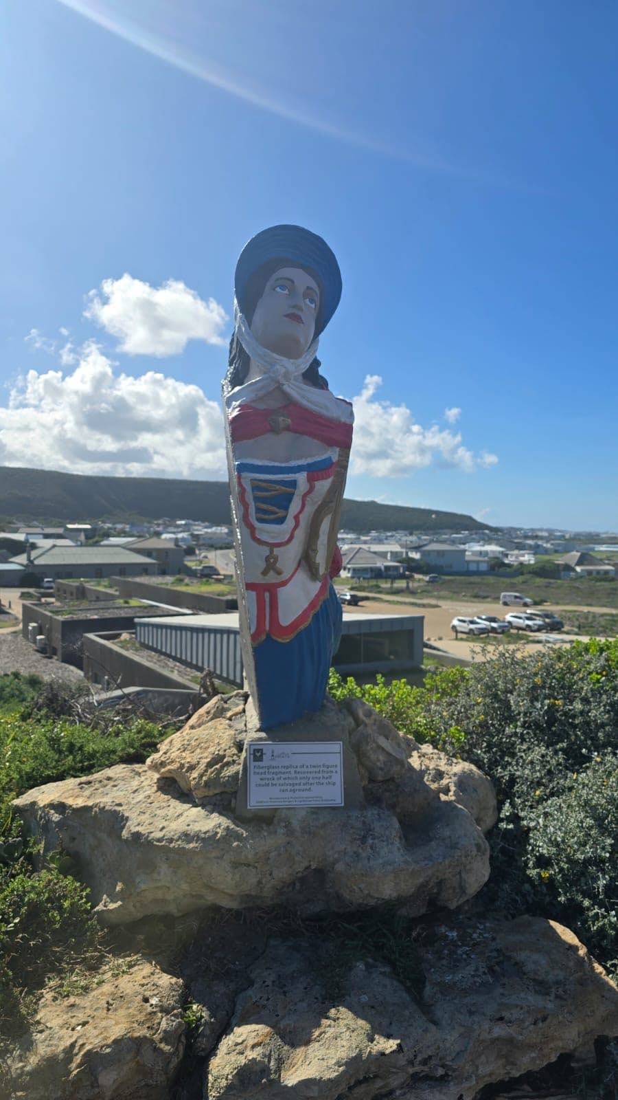 Colorful ship figurehead replica on a rocky pedestal overlooking a coastal town and hills.