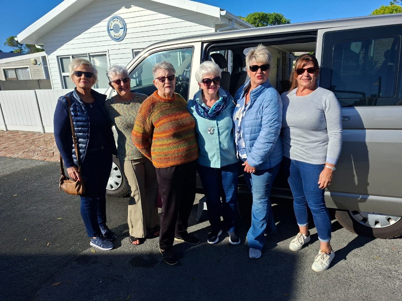 Six senior women wearing sunglasses and casual clothing pose together beside a silver passenger van.