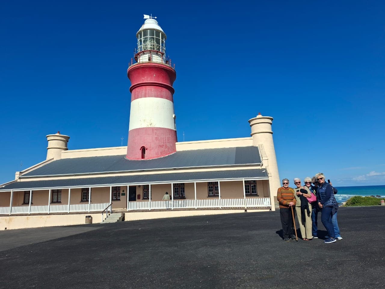 Red and white striped lighthouse and building with people posing under a clear blue sky.